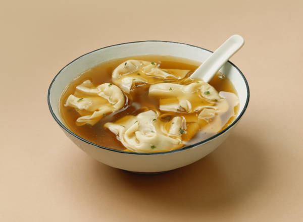 Eine Schüssel mit heißer Brühe, in der mehrere gefüllte Teigtaschen schwimmen. Die Teigtaschen sind hell und weich, und die Brühe hat eine goldene Farbe. Neben der Schüssel liegt ein Löffel. Der Hintergrund ist neutral und unauffällig.
