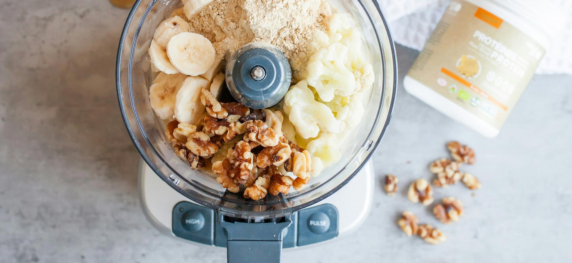 Ein Mixer enthält Bananen, Walnüsse, Blumenkohl und ein Proteinpulver. Die Zutaten sind gemischt und bereit zur Zubereitung eines gesunden Snacks oder Smoothies.