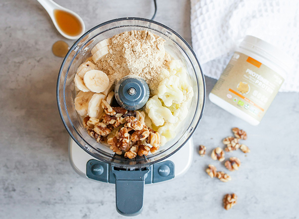 Ein Küchengerät ist mit Bananen, Blumenkohl, Walnüssen und Proteinpulver gefüllt. Neben dem Gerät liegen einige Walnüsse und ein Behälter mit dem Proteinpulver. Im Hintergrund befindet sich ein Löffel mit einem süßen Sirup.