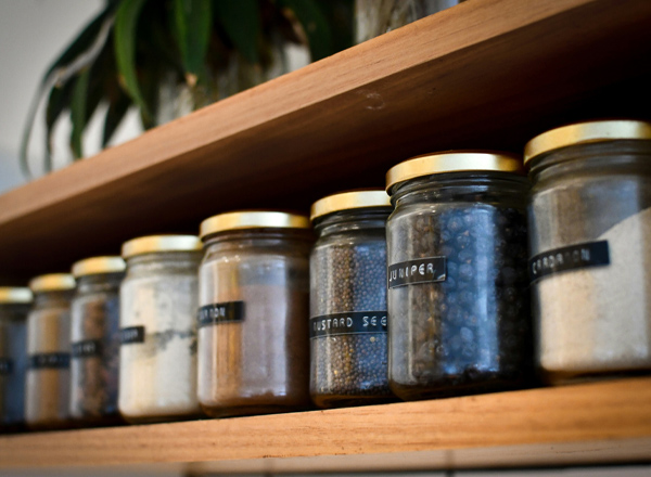 Auf einem Holzregal stehen mehrere Gläser mit verschiedenen Gewürzen und Samen. Jedes Glas hat ein Etikett, das den Inhalt beschreibt. Die Gläser sind beschriftet mit Namen wie „Juniper“ und „Mustard Seeds“. Im Hintergrund sind einige Pflanzen sichtbar.