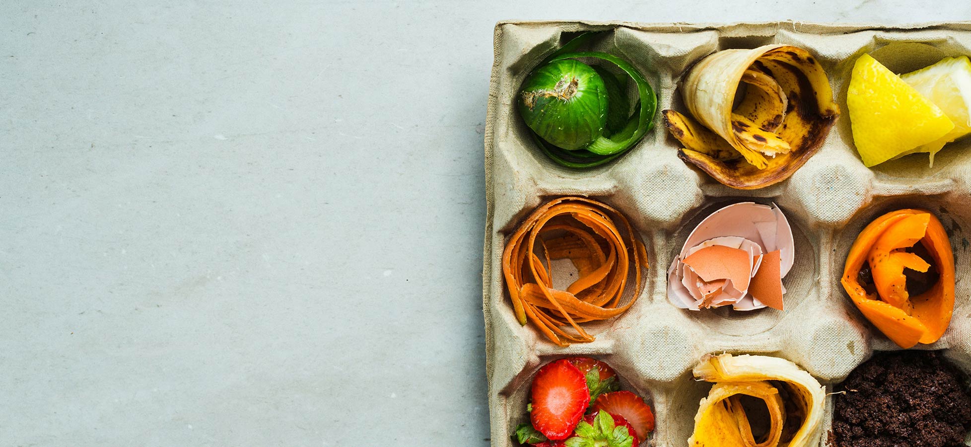 Eine Pappschachtel mit verschiedenen Küchenabfällen: Grünes Gemüse, Bananenschalen, Zitronenstücke, Karottenreste, Erdbeeren und Eierschalen sind angeordnet. Die Abfälle zeigen eine bunte Vielfalt an organischen Materialien, die zur Kompostierung bereit sind.