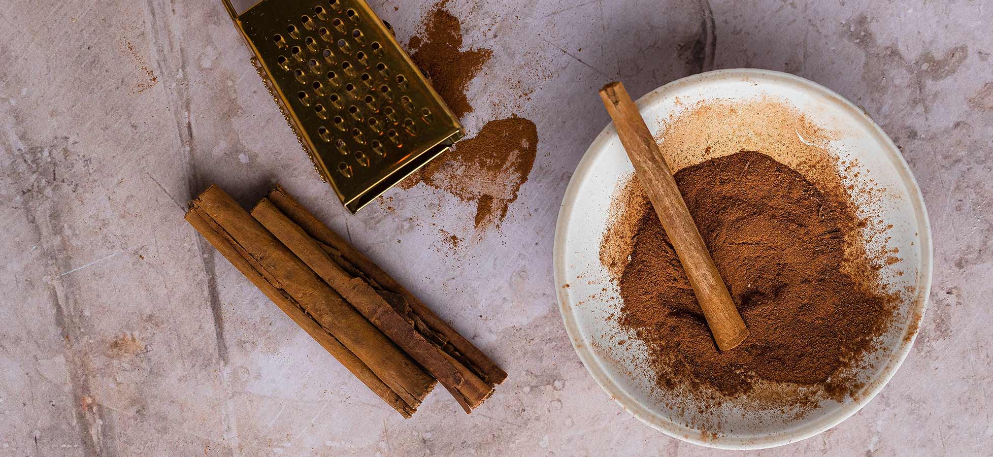 Eine Schüssel mit Zimtpulver und ein Zimtstock liegen auf einer Arbeitsfläche. Neben der Schüssel befindet sich eine goldene Reibe. Der Hintergrund ist grau marmoriert, was die natürlichen Farbtöne der Gewürze betont.