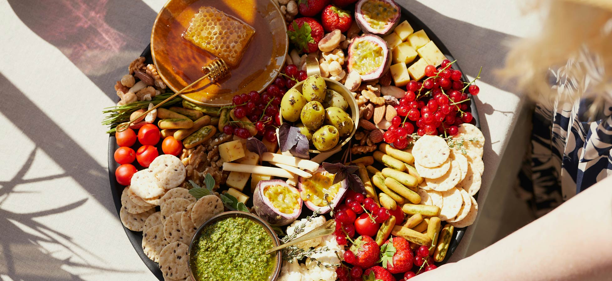 Ein runder Teller mit einer Vielfalt an Lebensmitteln: Käse, Früchte wie Erdbeeren und Kirschen, Oliven, Nüsse und Cracker. In der Mitte befindet sich ein Glas mit grünem Pesto und ein Stück Honig auf einem kleinen Teller. Hände greifen nach den Snacks.