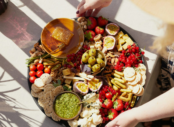 Ein runder Teller mit einer Vielfalt an Lebensmitteln: Käse, Früchte wie Erdbeeren und Kirschen, Oliven, Nüsse und Cracker. In der Mitte befindet sich ein Glas mit grünem Pesto und ein Stück Honig auf einem kleinen Teller. Hände greifen nach den Snacks.