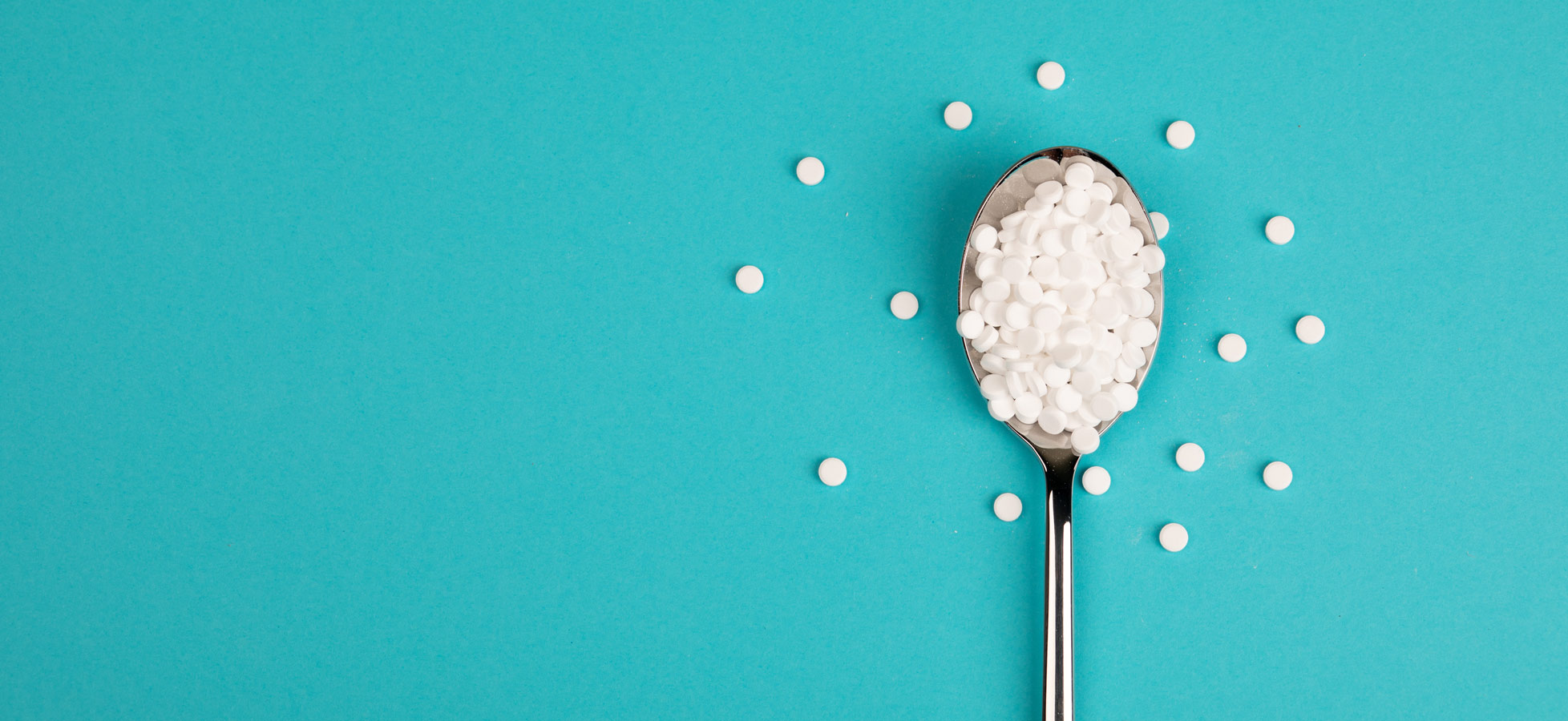 Ein Esslöffel mit weißen runden Pillen steht auf einem hellblauen Hintergrund. Neben dem Löffel liegen verstreut weitere ähnliche Pillen. Die Szene vermittelt den Eindruck von medizinischen oder homöopathischen Mitteln.