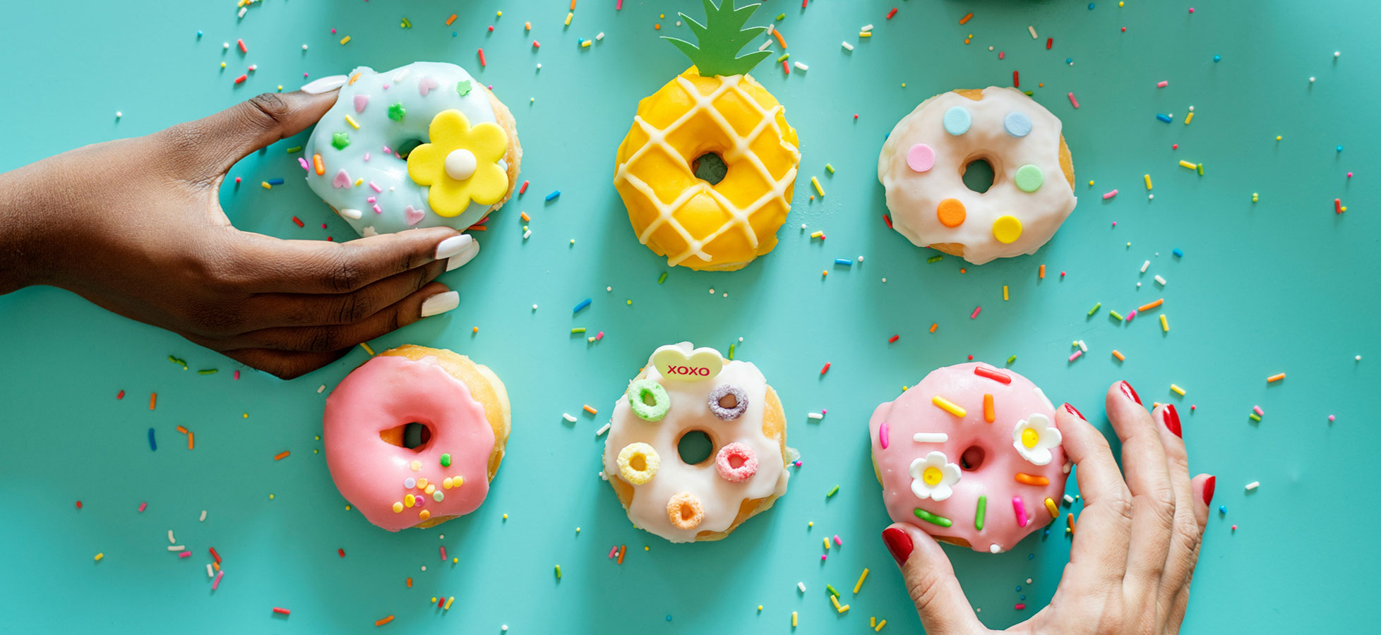 Sechs bunt dekorierte Donuts liegen auf einem hellblauen Tisch. Zwei Hände greifen nach den Donuts. Die Donuts sind in verschiedenen Farben und Formen, einige mit Streuseln und Zuckerguss verziert, darunter ein Donut in Ananasform. Bunte Streusel sind um die Donuts verstreut.