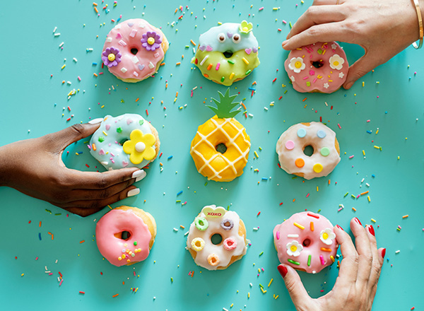 Auf einem hellblauen Tisch liegen bunte Donuts in verschiedenen Formen und Farben. Zwei Hände greifen nach den Donuts, die mit Blumen und Streuseln verziert sind. In der Mitte befindet sich ein Donut in Form einer Ananas. Der Tisch ist mit bunten Streuseln bedeckt.