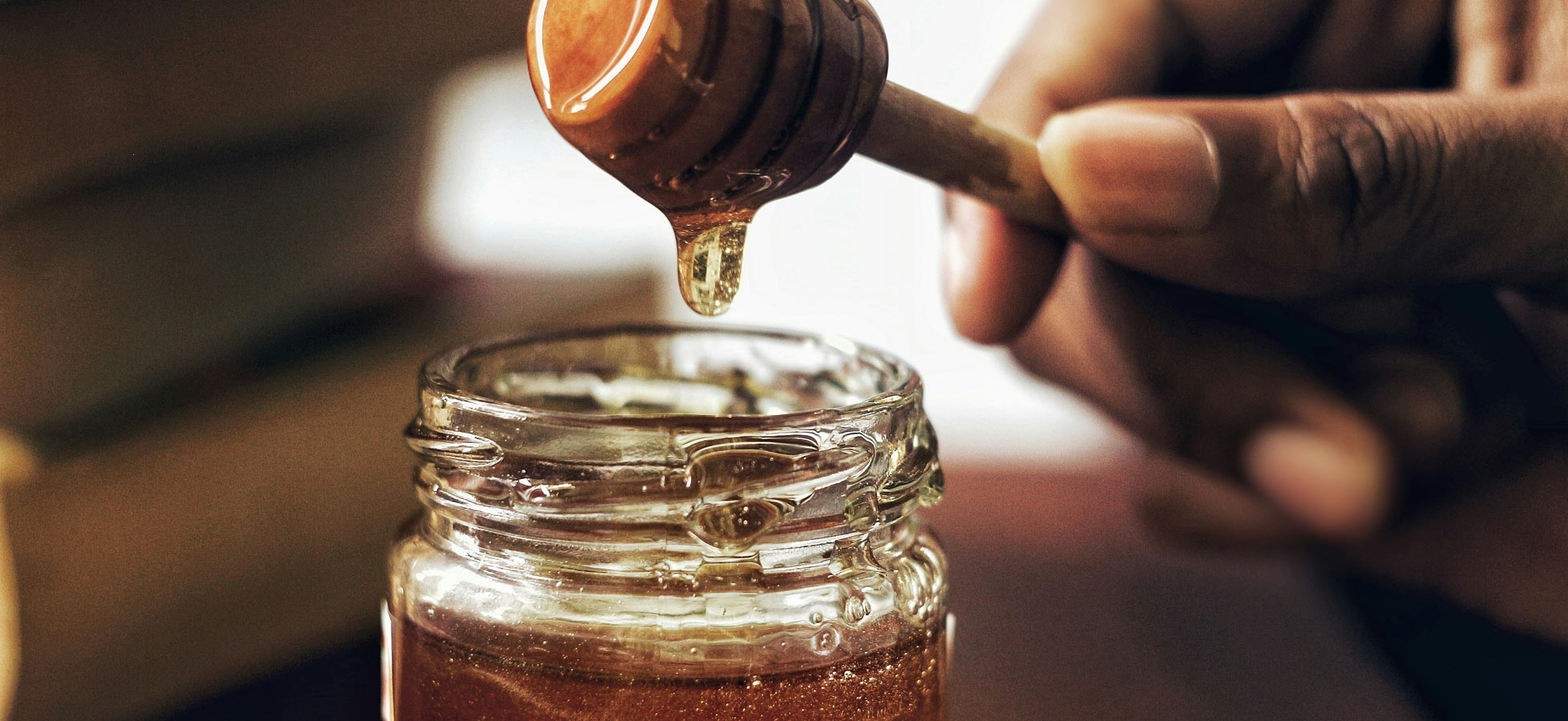 Ein Honiglöffel wird über ein Glas Honig gehalten, wobei der Honig langsam herabtröpfelt. Der Glasbehälter zeigt eine goldene, glänzende Flüssigkeit. Im Hintergrund sind unscharfe Umrisse von Bücher zu erkennen.