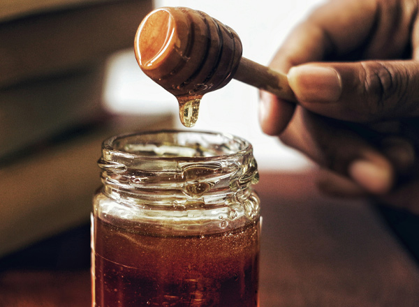 Ein Hand, die einen Honiglöffel hält, taucht in ein Glas Honig ein, aus dem der süße Honig tropft. Der Hintergrund ist unscharf, mit einem Hauch von Bücher.