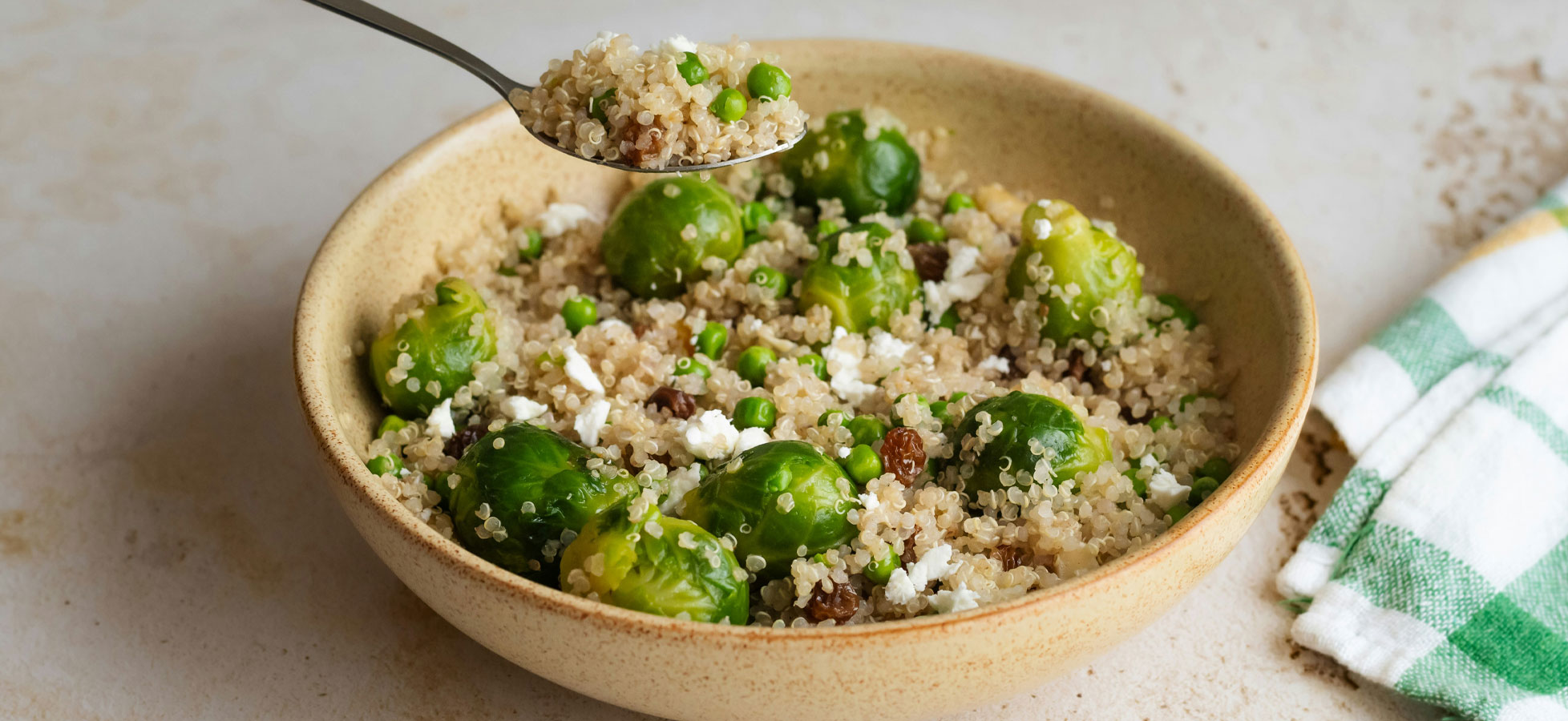 Eine Schüssel mit Quinoa, garniert mit grünen Erbsen und Rosenkohl. In der Schüssel sind auch kleine Stücke von Feta-Käse und Rosinen sichtbar. Ein Löffel hebt eine Portion der Mischung an.