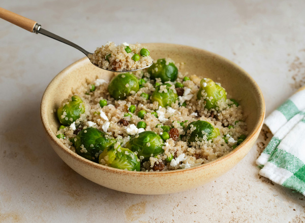 Eine Schüssel mit Quinoa, garniert mit grünen Erbsen und Rosenkohl. In der Schüssel sind auch kleine Stücke von Feta-Käse und Rosinen sichtbar. Ein Löffel hebt eine Portion der Mischung an.