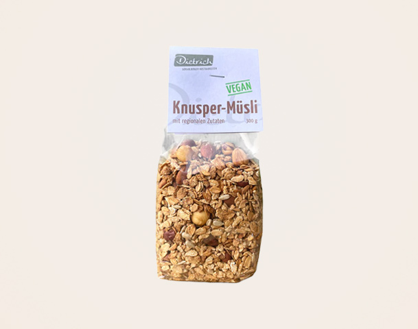 Ein transparentes Tütenpaket mit Knusper-Müsli. Die Verpackung ist beschriftet mit "Dietrich", "VEGAN" und "Knusper-Müsli", sowie dem Hinweis auf regionale Zutaten. Die Tüte enthält eine Mischung aus Haferflocken, Nüssen und anderen Müsli-Zutaten und wiegt 300 g.