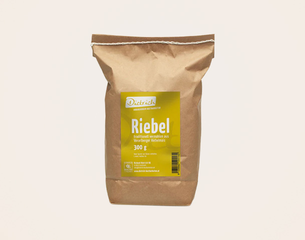 Das Bild zeigt eine braune Papierverpackung mit dem Aufdruck "Riebel" in gelber Schrift. Die Verpackung enthält 300 g Riebel, ein traditionelles Lebensmittel. Der Markenname "Dietrich" ist ebenfalls auffällig platziert.