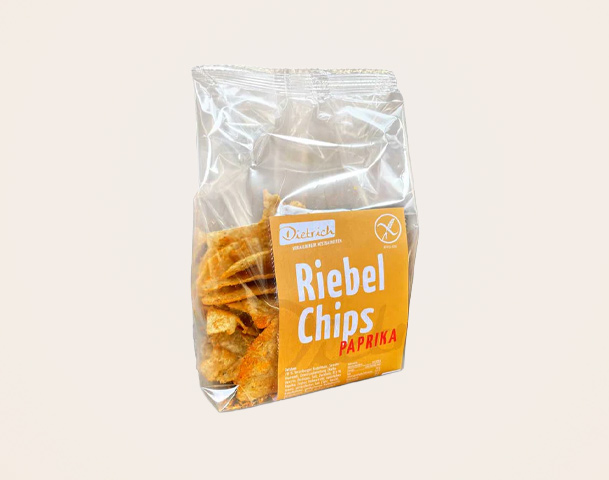 Eine transparente Tüte mit Riegel-Chips in Paprika-Geschmack. Die Verpackung trägt ein auffälliges Etikett mit dem Schriftzug "Riebels Chips Paprika" und dem Markennamen "Dettrich". Die Chips sind knusprig und goldbraun im Inneren der Tüte sichtbar.