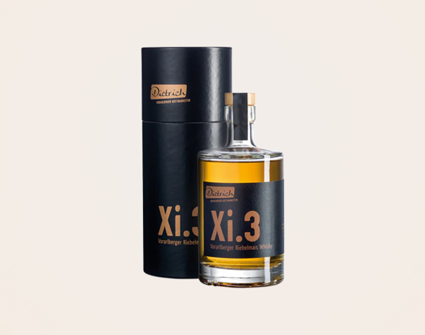 Eine Flasche Whisky mit dem Etikett „Xi.3“ steht neben einer schwarzen Verpackung. Der Whisky stammt aus Vorarlberg und ist in einer klaren Glasflasche mit einem Holzverschluss. Die Verpackung und die Flasche sind einfach und elegant gestaltet.
