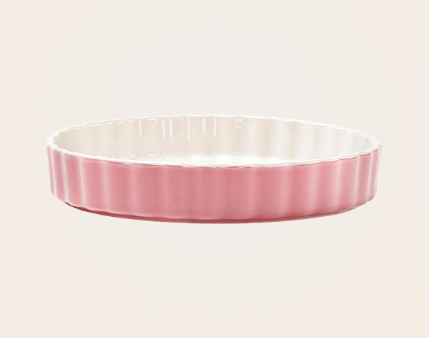 Eine runde, flache Auflaufform mit geriffeltem, rosa Rand und einer weißen Innenseite. Die Form hat eine glatte Oberfläche und eine elegante, schlichte Gestaltung, ideal für das Servieren von Speisen oder das Backen.