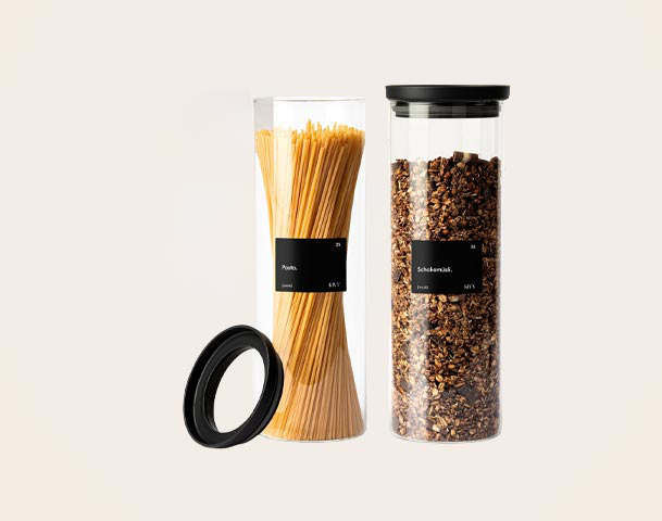 Das Bild zeigt verschiedene Vorratsbehälter. Ein hoher Behälter enthält Spaghetti, ein anderer ist mit Granola gefüllt. Daneben liegen Etiketten mit den Bezeichnungen "Weizenmehl" und "Pasta". Außerdem ist ein schwarzer Deckel zu sehen, der zu den Behältern passt.