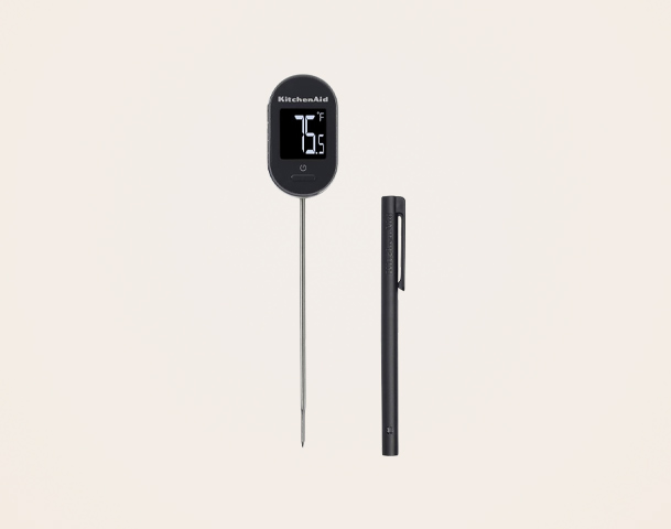 Das Bild zeigt ein digitales Küchenthermometer von KitchenAid mit einem langen Metallfühler, auf dem ein Display die Temperatur anzeigt. Neben dem Thermometer liegt ein schwarzer Stift. Der Hintergrund ist hell und unauffällig.