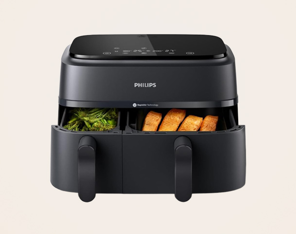 Ein schwarzer Heißluftfritteuse von Philips mit zwei Fächern. Im linken Fach sind grüne Brokkolistücke sichtbar, im rechten Fach liegen goldbraune, knusprige Lebensmittel. Die Oberseite hat ein digitales Bedienfeld mit Symbolen für verschiedene Funktionen.