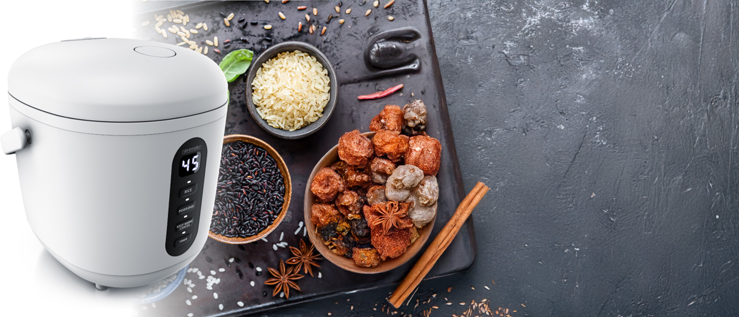 Ein Reiskocher steht links im Bild, daneben sind verschiedene Reisarten und asiatische Snacks in Schalen drapiert. Gewürze und Zutaten sind auf einer dunklen Oberfläche angeordnet. Der Reiskocher hat ein digitales Display mit der Temperaturanzeige.
