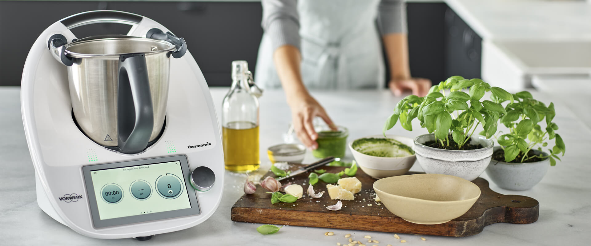 Ein Thermomix steht auf einem Tisch, während eine Person im Hintergrund frische Zutaten wie Basilikum, Knoblauch und Käse zubereitet. Der Thermomix hat einen Edelstahlbehälter und ein digitales Display, das Funktionen zur Zubereitung von Speisen anbietet.
