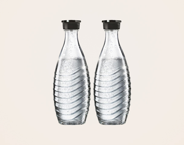Zwei klare Glasflaschen mit sprudelndem Wasser stehen nebeneinander auf einem hellen Hintergrund. Die Flaschen haben eine geschwungene Form und schwarze Deckel.
