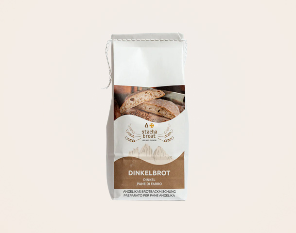 Eine Verpackung für Dinkelbrot mit dem Aufdruck „Dinkelbrot“ in großer Schrift. Darunter steht „Angelikas Brotbackmischung“ in kleinerer Schrift. Im Hintergrund sind Brotscheiben sichtbar. Die Verpackung hat eine weiße Grundfarbe mit braunen Akzenten.