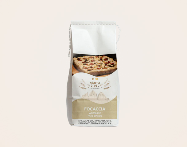 Eine weiße Papiertüte mit der Aufschrift „Focaccia Weißbrot“ und dem Logo „stacha brot“. Auf der Vorderseite ist ein Bild von Focaccia mit verschiedenen Toppings zu sehen. Die Tüte enthält eine Brotbackmischung.