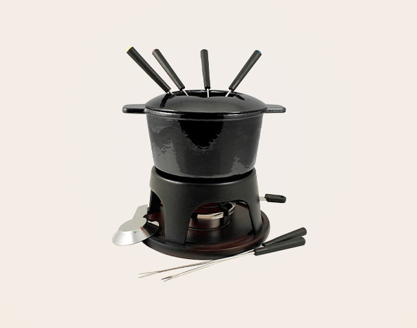 Ein schwarzes Fonduegerät steht auf einem stabilen Untergestell. Oben ist ein Topf mit fünf langen Gabeln, die hineinragen. Eine kleine Schale ist neben dem Gerät platziert. Das Gerät ist dafür gedacht, Speisen gemeinsam zu erwärmen und zu genießen.