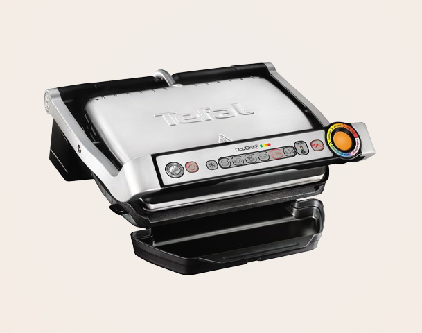 Ein Tefal-Kontaktgrill mit silbernem Gehäuse und schwarzen Bedienelementen. Die Grillplatte ist oben aufgeklappt und zeigt eine saubere Grillfläche. An der Vorderseite befindet sich ein farbiger Drehregler sowie mehrere Tasten zur Temperatureinstellung und für verschiedene Grillfunktionen.
