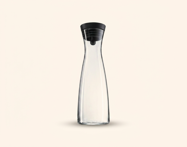 Eine klare Glas-Wasserkanne mit einem schwarzen Deckel steht auf einem hellen Hintergrund. Die Kanne hat eine elegante, schlanke Form und ist nach oben hin leicht verjüngt.