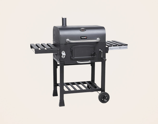 Ein schwarzer Grill mit zwei seitlichen Ablageflächen und einem Untergestell auf Rollen. Der Grill hat eine geschlossene Kammer mit einer Tür und einem Kamin auf der Oberseite. Die Konstruktion ist stabil und eignet sich für das Grillen im Freien.