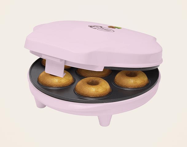 Ein rosa Donut-Maker mit geöffnetem Deckel, der fünf frisch gebackene Donuts zeigt. Die Donuts sind goldbraun und formenrund. Der Gerät hat eine einfache, kompakte Form und steht auf kleinen Füßen.