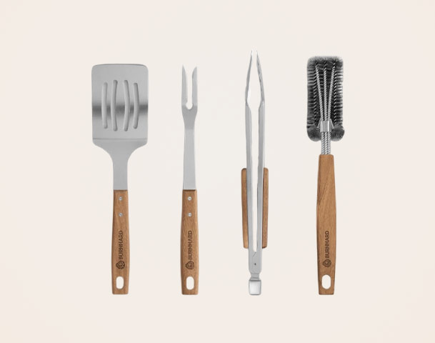 Eine Grillbesteck-Set bestehend aus vier Werkzeugen: einem Wender, einer Gabel, einer Zange und einer Bürste. Alle Werkzeuge haben Edelstahlköpfe und Holzgriffe, die für einen besseren Halt sorgen. Sie sind auf einem einfarbigen Hintergrund angeordnet.