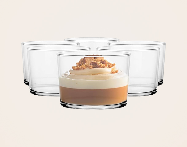 Auf dem Bild sind fünf klare Gläser zu sehen, von denen eines im Vordergrund steht. Dieses Glas enthält eine Schicht aus karamellfarbener Creme, gekrönt mit einer Schicht Sahne und Keksbruch. Die restlichen Gläser stehen im Hintergrund und sind leer.