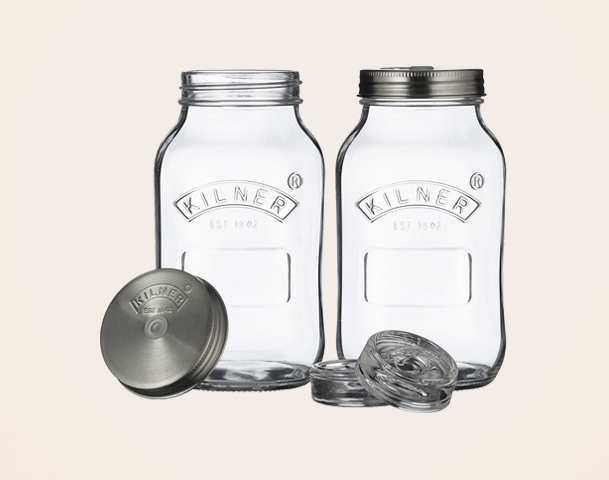Zwei gläserne Einmachgläser von Kilner stehen nebeneinander. Eines hat einen metallenen Schraubdeckel, das andere einen klassischen Deckel. Zusätzlich liegen weitere Deckel auf dem Tisch. Die Gläser sind für die Konservierung von Lebensmitteln geeignet.