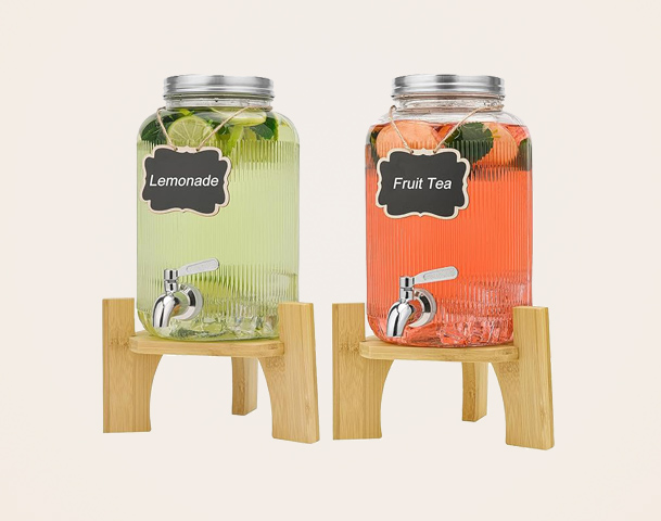 Zwei Glasbehälter stehen nebeneinander auf einem Holzständer. Der linke Behälter enthält grüne Limonade mit Limetten, der rechte fruchtigen Tee mit verschiedenen Früchten. Beide Behälter haben einen Auslass zum Zapfen der Getränke und sind mit kleinen Schildern beschriftet.