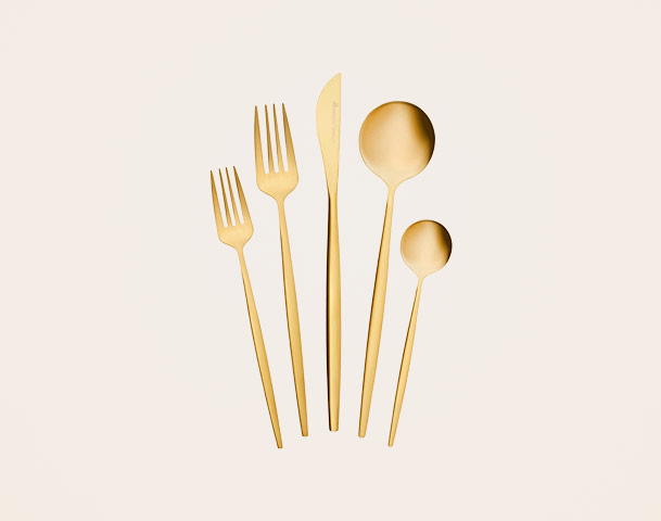 Eine Gruppe goldfarbener Bestecke liegt auf einem hellen Hintergrund: zwei Gabeln, ein Messer und zwei Löffel in verschiedenen Größen. Das Design ist modern und schlicht.