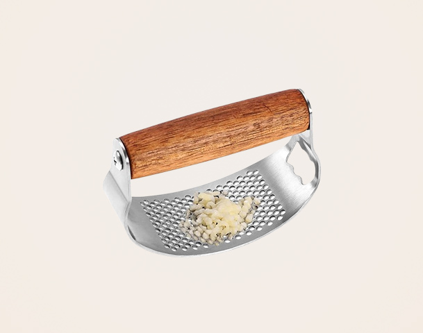 Ein Knoblauchzerkleinerer mit einem Holzgriff aus Edelstahl, auf dem feingehackter Knoblauch liegt. Im Hintergrund sind ganze Knoblauchzehen und ein Reinigungswerkzeug sichtbar. Das Bild zeigt alles, was für die Zubereitung von Knoblauch benötigt wird.