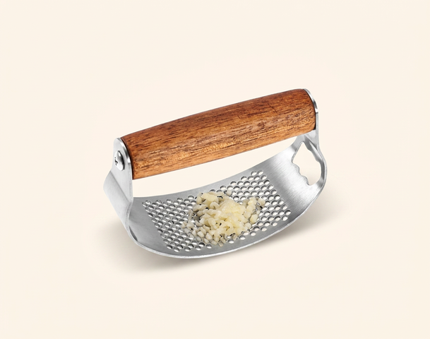 Ein Knoblauchzerkleinerer mit einem Holzgriff aus Edelstahl, auf dem feingehackter Knoblauch liegt. Im Hintergrund sind ganze Knoblauchzehen und ein Reinigungswerkzeug sichtbar. Das Bild zeigt alles, was für die Zubereitung von Knoblauch benötigt wird.