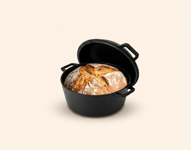 Ein gusseiserner Topf mit frisch gebackenem Brot, das eine goldene Kruste hat, steht im Vordergrund. Im Hintergrund sind eine Schachtel mit Rezepten sowie einige Weizenkörner zu sehen. Der Fokus liegt auf dem Brot und dem Topf.