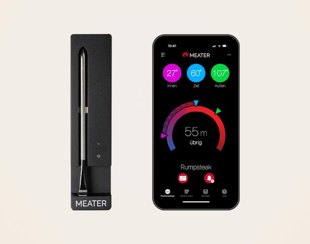 Das Bild zeigt ein kabelloses Fleischthermometer namens MEATER, das in einer Halterung steht, sowie ein Smartphone mit einer App. Die App zeigt Temperaturen für Innen, Ziel und Außen sowie die verbleibende Garzeit für ein Rumpsteak an.