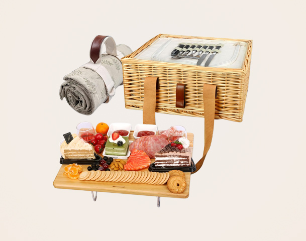 Ein Picknickkorb aus Korbgeflecht mit einem gedeckten Holzbrett. Auf dem Brett sind verschiedene Snacks angeordnet, darunter Salami, Obst, Kuchen und Kekse. Eine Decke liegt neben dem Korb, der hübsch mit Besteck und Servietten gefüllt ist.