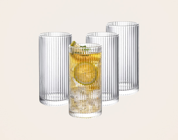 Vier transparente Gläser mit einem gerippten Design. In der Mitte steht ein Glas mit einem hellbraunen Getränk, garniert mit einer Zitronenscheibe und einem Limettenstück. Die anderen Gläser sind leer und stehen daneben.