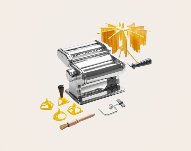 Eine klassische Nudelmaschine aus Edelstahl steht im Mittelpunkt, umgeben von verschiedenen Nudelausstechformen, einem Pinsel und einem Halter für frische Pasta. Die Maschine hat einen Handkurbelmechanismus, um den Teig zu walzen.