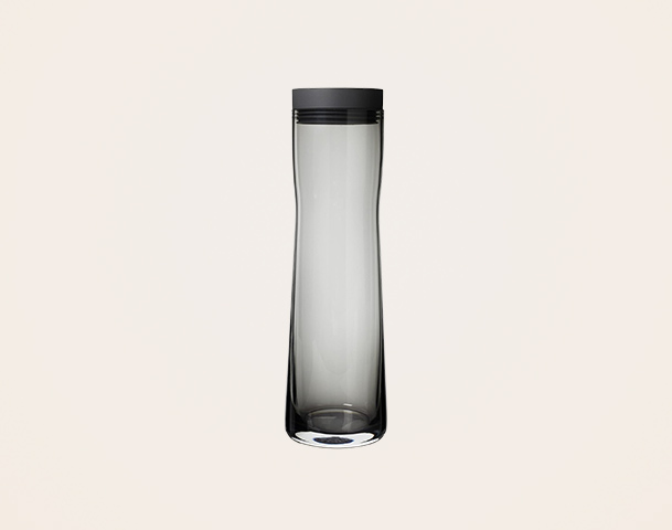 Eine schlanke, gläserne Wasserflasche mit einem schwarzen Deckel. Die Flasche ist transparent und zeigt einen klaren Blick auf den Inhalt, mit einer leicht geschwungenen Form, die an der Basis breiter wird. Ideal für die Aufbewahrung von Getränken.
