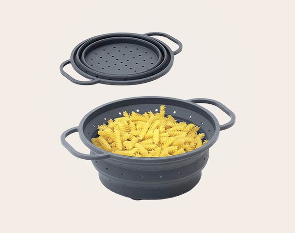 Eine grau silikonisierte Abtropfschale mit langen Griffe, die mit ungekochten Pasta gefüllt ist. Darüber liegt ein passender Deckel mit Löchern für Dampfdurchlässigkeit. Die Schale ist flexibel und dient zum Abtropfen oder Dämpfen von Lebensmitteln.
