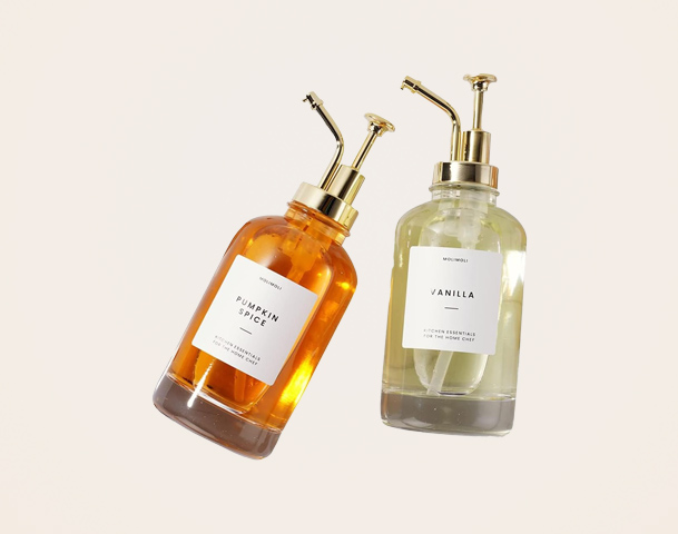 Zwei elegante Flaschen mit Pumpaufsätzen, eine orange mit der Beschriftung „Pumpkin Spice“ und eine transparente mit der Aufschrift „Vanilla“. Beide Flaschen haben goldene Details und stehen vor einem hellen Hintergrund. Die Produkte scheinen für Raumdüfte oder ätherische Öle gedacht zu sein.