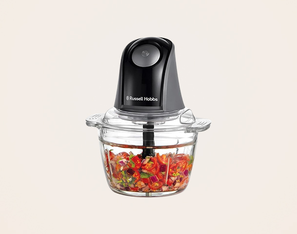 Ein schwarzer Zerkleinerer von Russell Hobbs steht über einer klaren Schüssel, die mit klein geschnittenem Gemüse gefüllt ist. Der Zerkleinerer hat einen runden Knopf an der Oberseite und zeigt, dass er zum Zerkleinern von Zutaten verwendet werden kann.