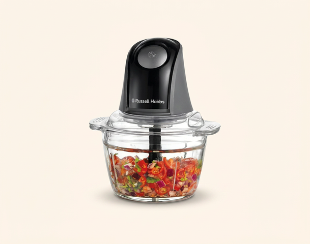 Ein schwarzer Zerkleinerer von Russell Hobbs steht über einer klaren Schüssel, die mit klein geschnittenem Gemüse gefüllt ist. Der Zerkleinerer hat einen runden Knopf an der Oberseite und zeigt, dass er zum Zerkleinern von Zutaten verwendet werden kann.
